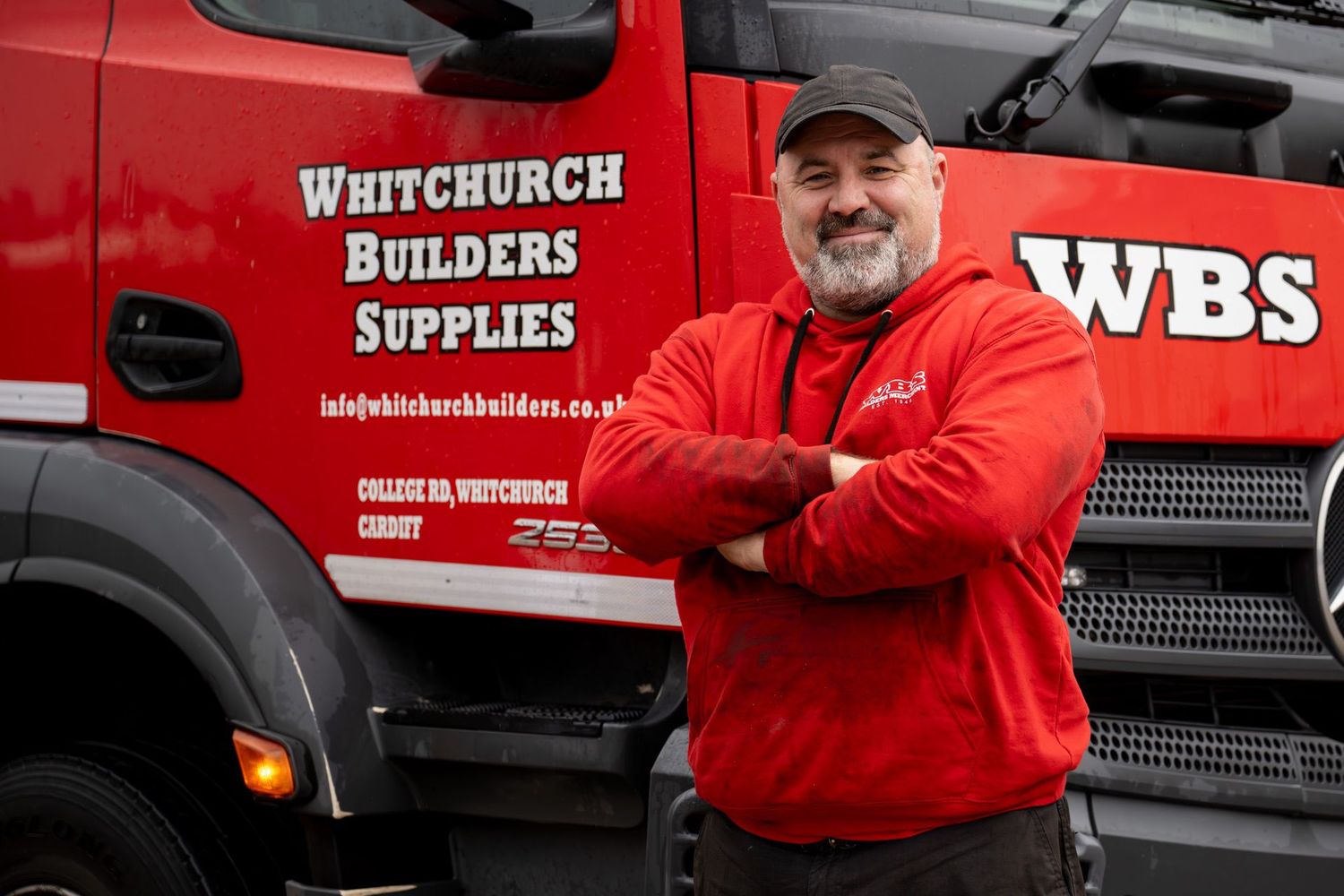 A smiling man in a red uniform with crossed arms standing in front of a red WBS Builders truck.