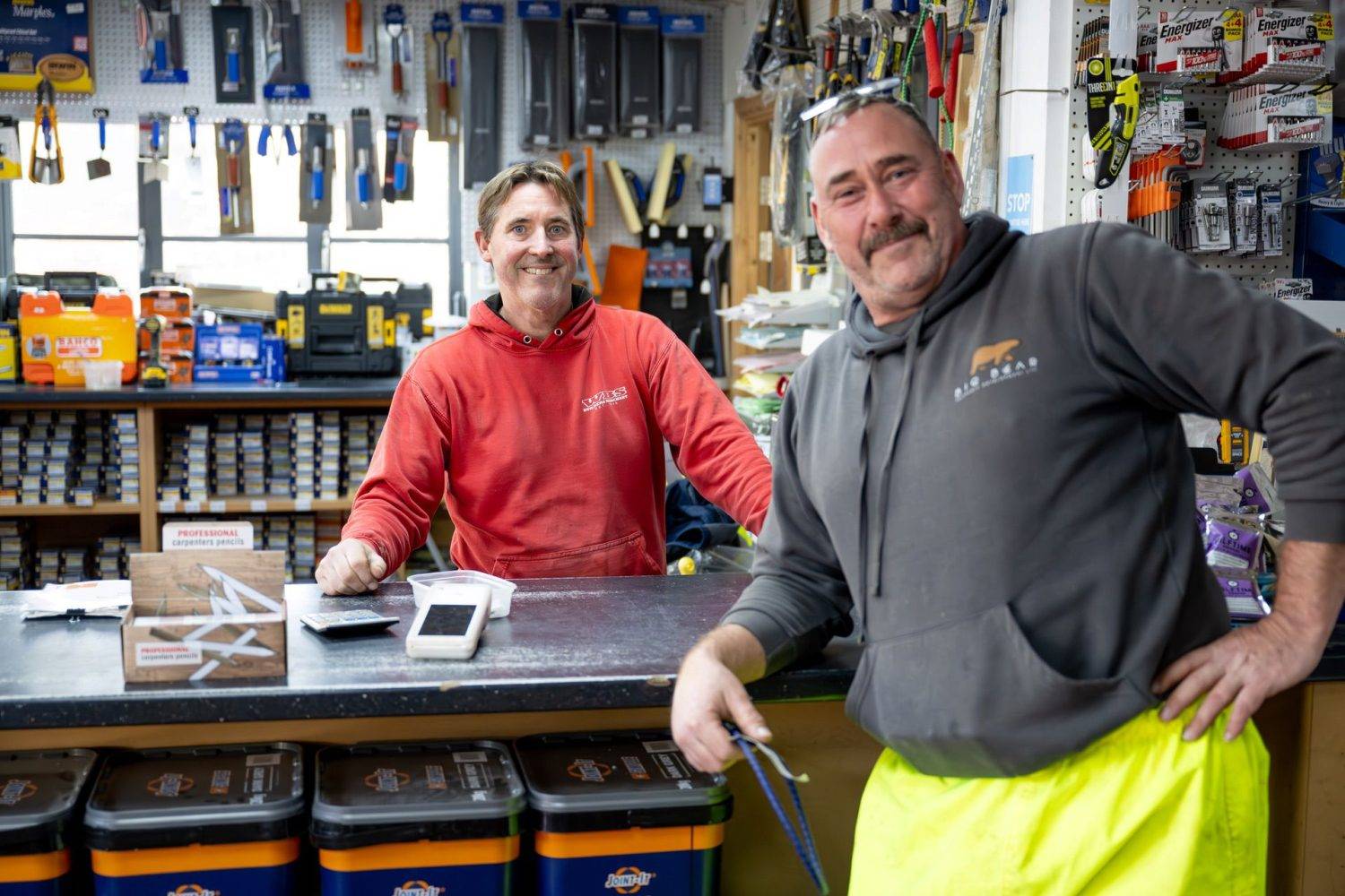 Two men smiling at the camera, one in a red sweatshirt and the other in a gray hoodie, standing behind a counter in builders' merchants store.