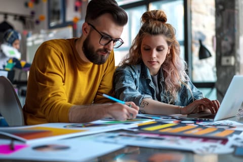 Two individuals sitting at a table are collaborating on an art project, with one drawing on paper and the other designing a website on their laptop. The table is covered with various art materials and papers, blending traditional creativity with modern e-commerce tips.