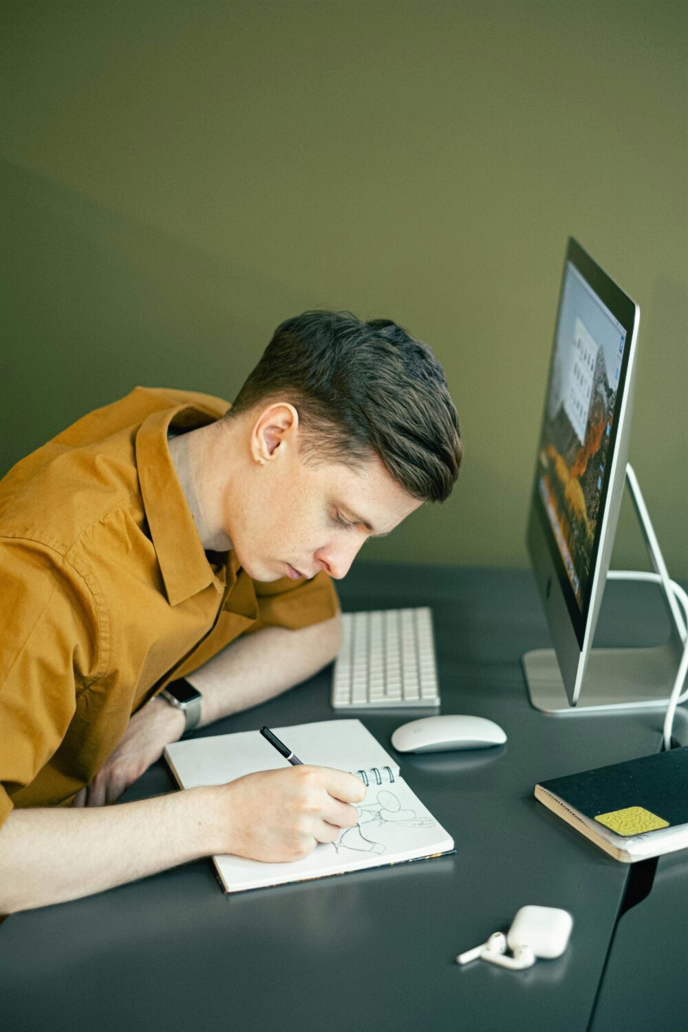 Person in a yellow shirt sitting at a desk, drawing in a notebook with an iMac computer, keyboard, and mouse in front of them, possibly brainstorming best practices for creating a user-friendly website.