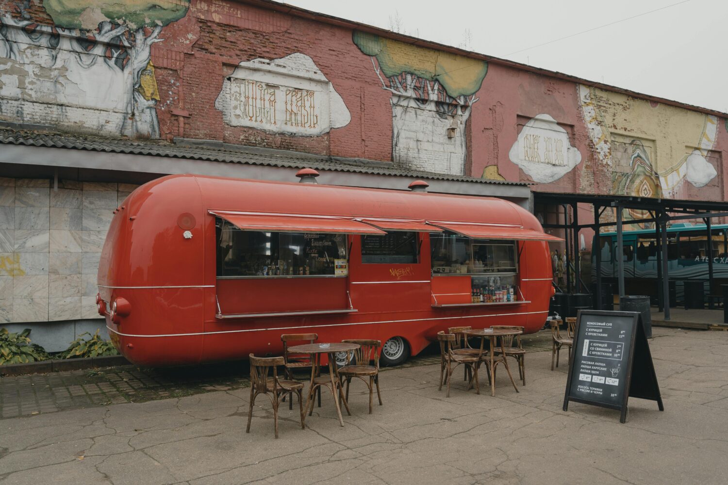 A vibrant red food truck with open windows is stationed by a weathered mural. In front, three wooden tables with chairs invite diners, while a chalkboard menu showcases the day's offerings. Photos of this scene enhance the allure on the restaurant's website, optimizing SEO and featuring downloadable PDF menus.